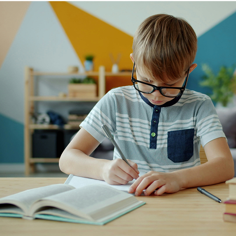 A young boy with glasses, seated at a table, writing in a notebook in a homeschool setting.