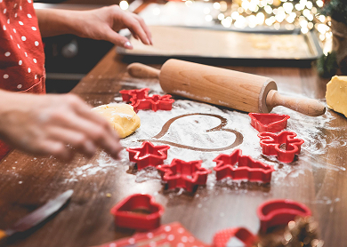 a persons hands at a countertop with a rolling pin, christmas shaped cookie cutters and flour sprinkled in the center with a heart drawn into it 
