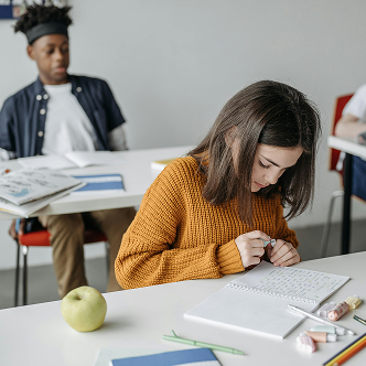 A classroom with a girl sitting in front, writing in her notbook, and a boy sitting behind her, leaning back in his chair looking down at the books on his desk.