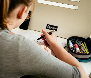 An over the shoulder view of a girl doing her homework with an open pencil case next to her.